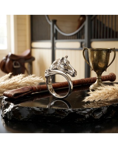 Bague argent chevalier en forme de tête de cheval, sur socle noir, près d’une coupe dorée et d’un fleuron en paille, ambiance éq