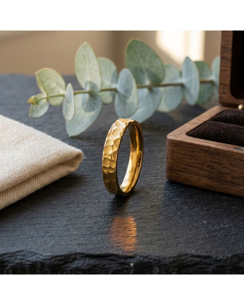 Alliance demi-martelée homme en or posé sur une plaque sombre, avec boîte en bois et brindille de eucalyptus en arrière-plan.