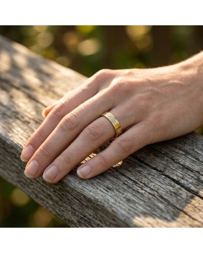Alliance effet bois femme en or : doigt portant une bague dorée gravée, posé sur une planche en bois.