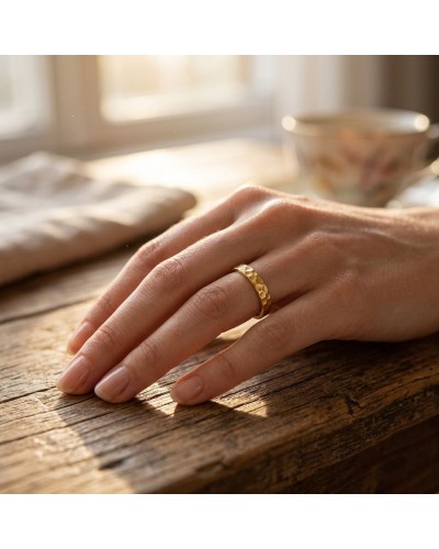 Alliance irrégulière martelée femme en or portée sur le doigt, posé sur une table en bois avec une tasse en porcelaine au fond.