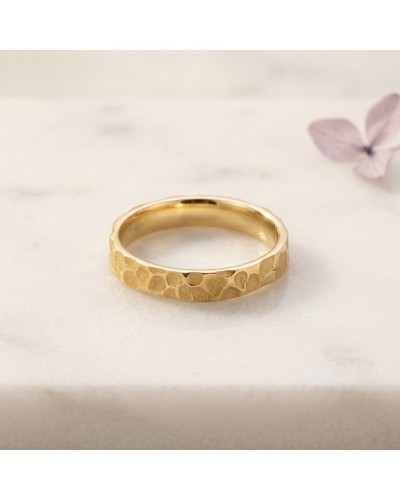 Alliance irrégulière martelée femme en or: bague dorée texturée posée sur surface claire avec fleur violette en arrière-plan