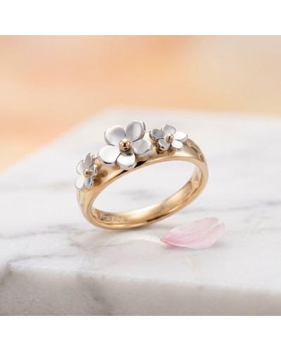 Bague dorée Cerisier avec trois fleurs argentées et petites billes au centre, sur fond marbre et pétale rose posé à côté.