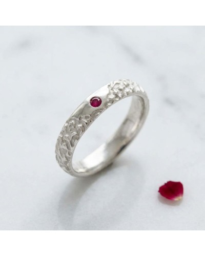 Alliance BISON homme en or: bague argentée texturée avec petite gemme rouge, posé sur surface claire avec pétale rouge nearby