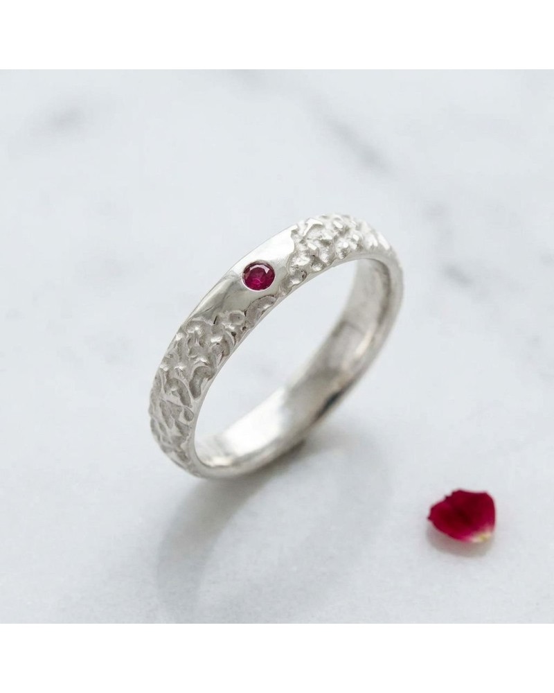 Alliance BISON homme en or: bague argentée texturée avec petite gemme rouge, posé sur surface claire avec pétale rouge nearby