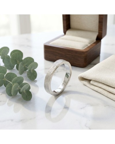 Alliance NERVURE 4 mm Homme en or: alliance argentée à nervures texturées posée sur marbre, bijoux et coffret en bois en arrière