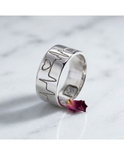 Alliance BEATING LOVE homme en or: bague argentée gravée avec cœur, posée sur marbre blanc, pétale rose au pied.