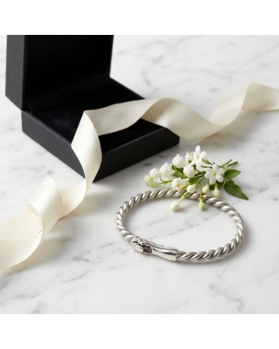 Bracelet en argent rhodié torsadé posé près d’un ruban blanc et de petites fleurs blanches, présent dans une boîte noire.