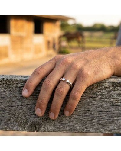 Bague TROT homme en argent rhodié sur le doigt d’un homme posé sur une clôture en bois, fond champ et écuries floues.