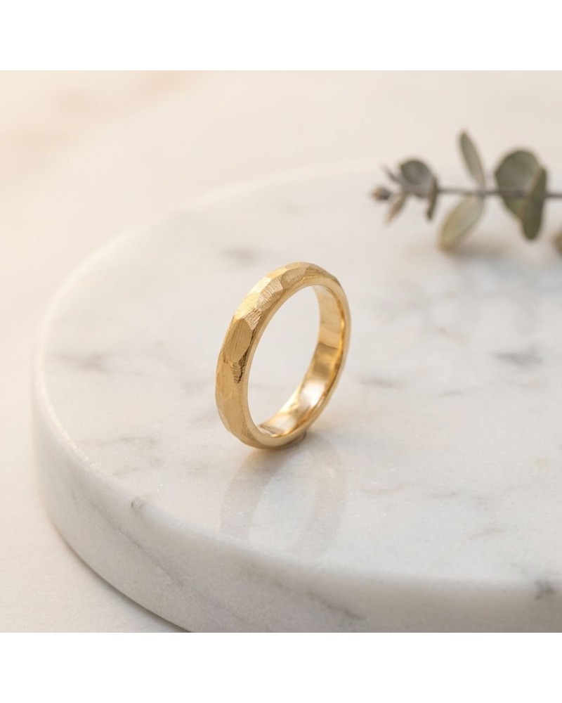 Alliance FLASH 4 mm femme en or: bague dorée à surface texturée sur socle en marbre, fond flou, petite branche verte en arrière-