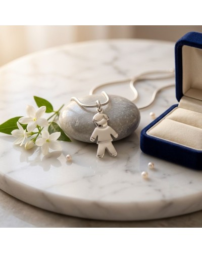 Pendentif en forme d'enfant argenté, sur chaîne, posé sur une pierre gris, près d’un boîtier en velours bleu et fleurs blanches.