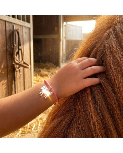 Bracelet léger en cordon rose avec pendentif cheval argenté sur le poignet d’un enfant, dans une étable au fond de laquelle on v