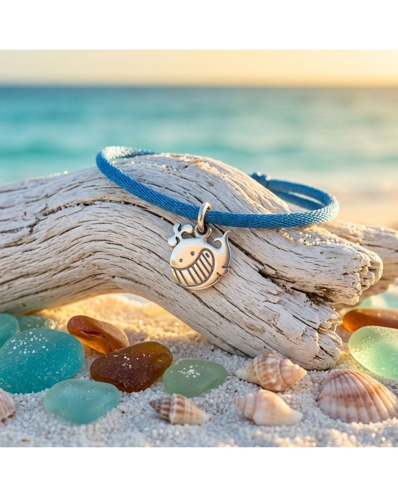 Bracelet bleu en cordon avec pendentif fantaisie en forme de baleine souriante sur bois flotté, plage et coquillages au soleil.