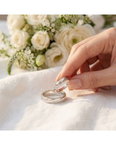 Alliance entrelacée en argent avec bouquet de fleurs blanches en arrière-plan, posé sur lin.