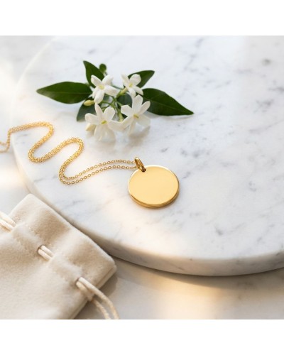 Médaille jeton en or jaune 18k sur chaîne fine, posée sur plateau de marbre avec petites fleurs blanches en arrière-plan.