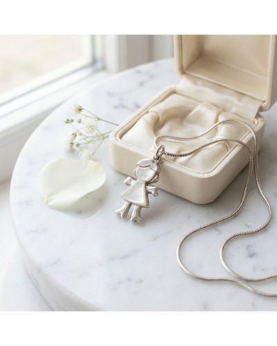 Pendentif EVE en forme de petite fille argentée, sur une chaîne serpentine, posé près d’une boîte à bijoux beige et d’une pétale