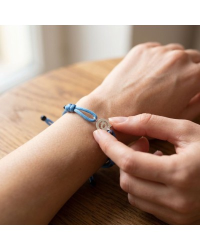 Bracelet bleu à cordons ajustables autour du poignet, plaquette métallique ronde avec symbole, portant le nom de produit. Bracel