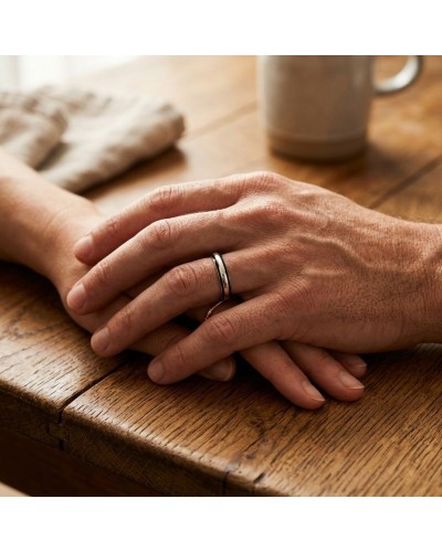 Alliance MODESTE Homme en or: deux mains jointes sur une table en bois, porte une bague masculine argentée et noire.