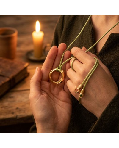Pendentif Anneau Zoom en or matté porté par mains entouré de corde vert olive, candle et livre en arrière-plan.