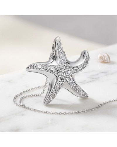 Pendentif étoile de mer argenté orné de cristaux, collier en chaîne délicate posé sur marbre, coquillage à côté.