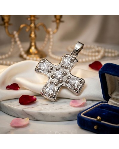 Pendentif croix baroque en argenté orné de brillants, posé sur satin blanc avec pétales roses, casier bleu en arrière-plan. incl