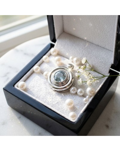 Pendentif BOUEE argenté avec pierre centrale bleue dans boîte noire, entouré de perles et petites fleurs blanches.