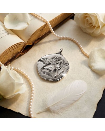 Pendentif rond argenté gravé d’un ange ailé, posé près d’un livre ouvert et d’une perle, sur une plume blanche.