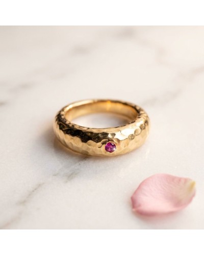 Anneau en or martelé avec pierre rose violet (DEM JONC) posé sur une surface claire, pétale de rose à côté.