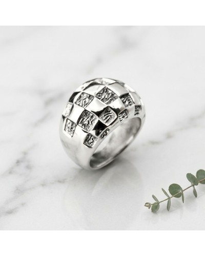 Bague DAMIER argentée à motifs damier 3D sur fond blanc, surface brillante avec reflets, près d’une petite branche.