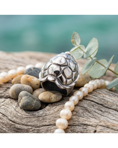 Bague TORTUE argentée avec motif géométrique, posée sur pierre et bois, accompagnée de perles et de verdure.