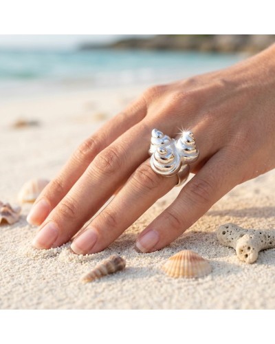 Anneau argenté en forme de coquillages porté sur le doigt, posé sur le sable près de la mer avec des coquilles et coraux en déco