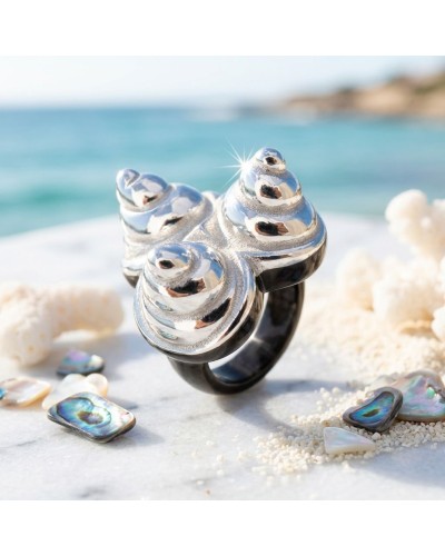 Bague COQUILLAGES en argent miroir avec motif spirale, posée sur sable près de la mer, coquillages colorés autour.