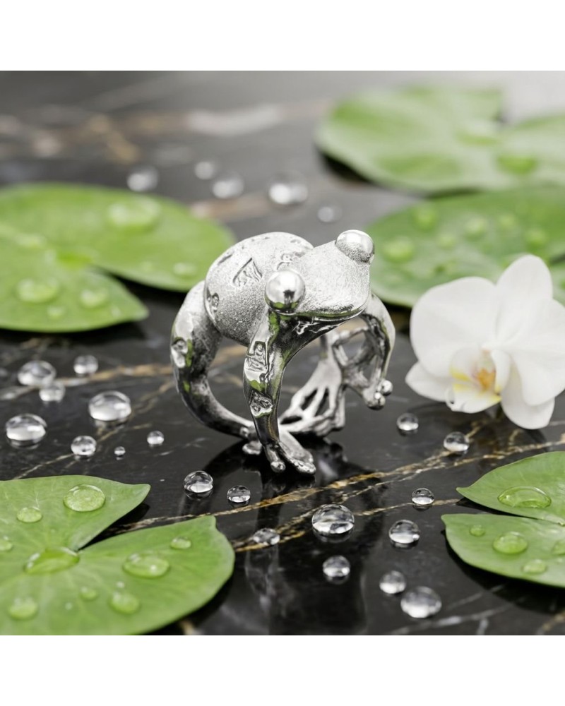 Bague argentée en forme de grenouille sur une surface noire, entourée de feuilles et d’une fleur blanche, avec des gouttes d’eau