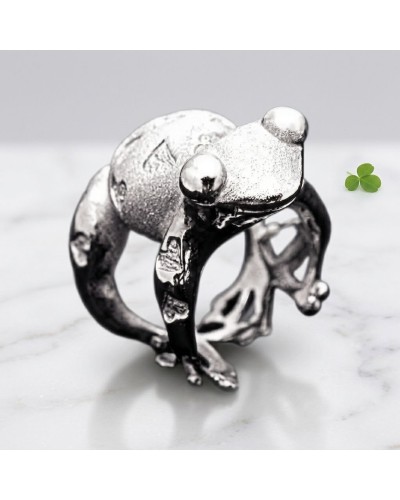 Bague argentée en forme de grenouille, détail peau texturée, pose sur marbre avec petit trèfle vert à côté.