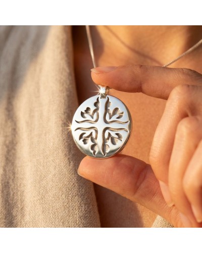 Pendentif MATAHARI circulaire argenté avec motif feuille d’arbre découpé, posé entre les doigts.