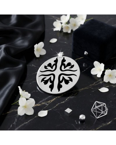 Pendentif rond argenté gravé d’un motif noir stylisé en forme de croix florale, posé sur marbre noir avec fleurs blanches.