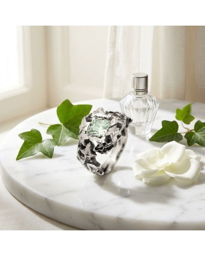 Bague LIERRE en argent avec motif feuilles et pierre centrale verde, posée sur table marbre avec feuilles et parfum.