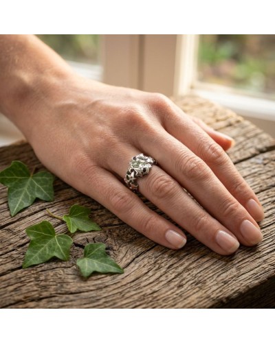 Bague LIERRE en argent portée sur le doigt, motifs feuillagés et tête de sangle, posée sur bois avec feuilles vertes autour.