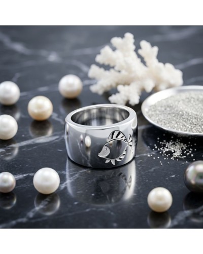 Bague argentée avec motif poisson IKAN gravé, posée sur surface sombre entourée de perles et de décorations marines.
