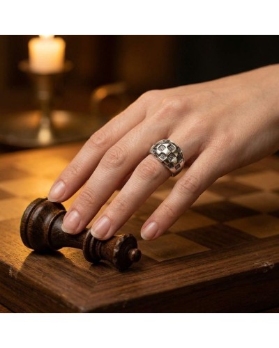 Bague DAMIER en argent avec motif damier, portée sur l’annulaire d’une main posé sur une pièce d’échecs en bois.