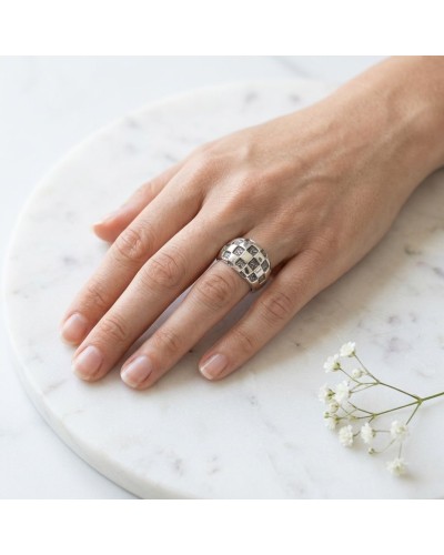 Bague DAMIER argentée à motif damier sur le doigt, posé sur une surface marbrée avec petites fleurs.
