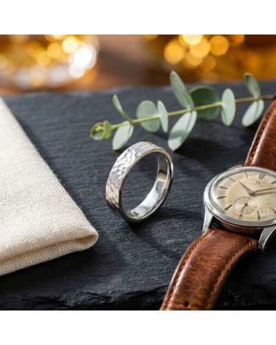 Alliance homme en alliage martelé argenté posé à côté d'une montre à bracelet cuir et d'un brin de feuilles, ambiance élégante.