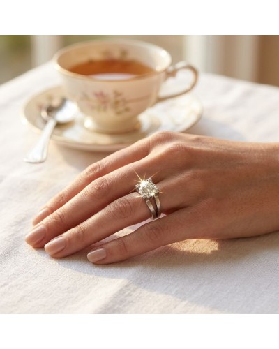 Bague ROMANTIS sur doigt, pierre centrale brillante avec double bande argentée, près d’une tasse de thé sur nappe blanche.