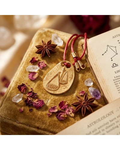 Pendentif zodiaque Balance en or avec cordon rouge et fermoir offert, posé sur livre et décorations astrologiques.