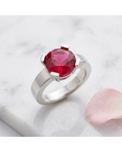 Bague argentée avec pierre ronde rouge vif, sertissure à quatre griffes élégantes, sur fond marbré. Produit Bague HASRAT.