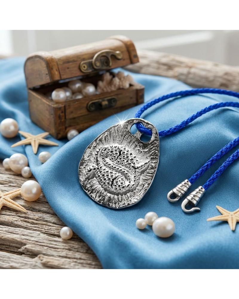 Pendentif zodiaque Poisson en métal texturé avec cordon bleu et fermoir offert, posé sur tissu bleu près d’un coffre et perles.