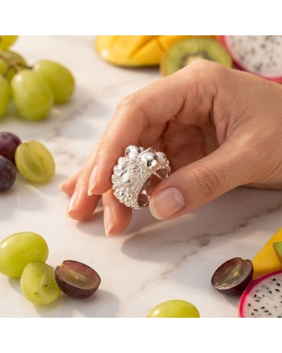 Bague argentée ornée de motifs floraux tenue entre les doigts, fruits tranchés et grappes de raisin autour.