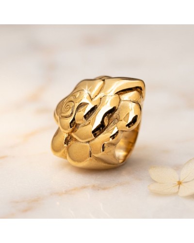 Bague dorée somptueuse en métal brillant avec motifs incurvés, posé sur une surface claire, petite fleur blanche à côté. Mot clé