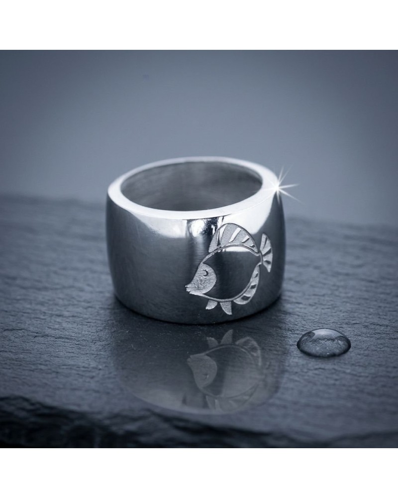 Bague épaisse en métal argenté avec gravure poisson, reflet sur une surface sombre et perle d’eau à côté. (Bague IKAN)