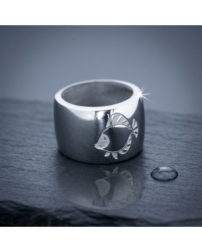 Bague épaisse en métal argenté avec gravure poisson, reflet sur une surface sombre et perle d’eau à côté. (Bague IKAN)