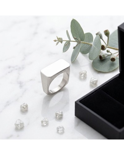 Anneau argenté moderne à forme rectangulaire « Bague CLEAN RECTANGLE » posé sur marbre avec pierres et branche d’eucalyptus.